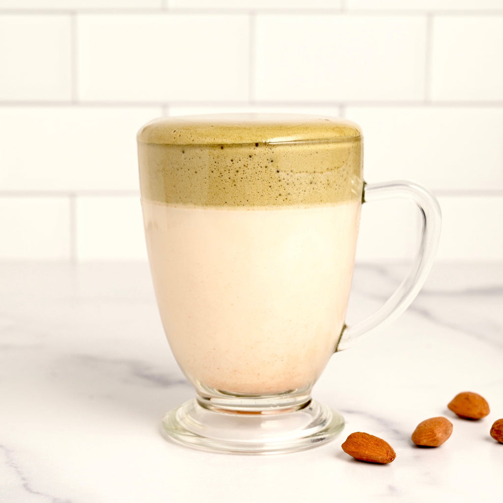A clear glass latte mug is filled with a layered hot beverage, the bottom layer is white and the top layer is green, a few almonds are scattered near the mug
