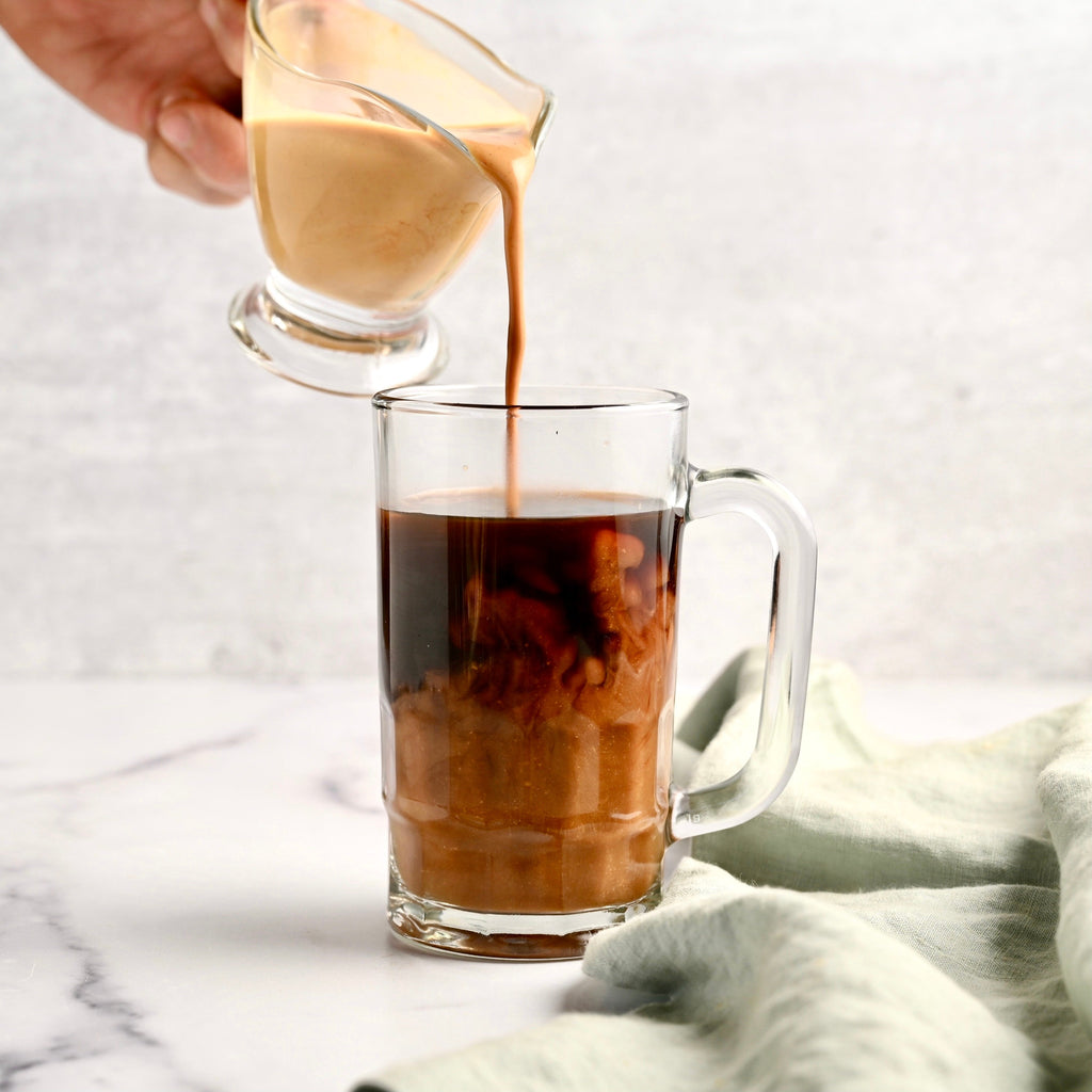 A clear glass coffee mug contains dark roasted coffee as a hand pours gingerbread creamer from another clear glass cup