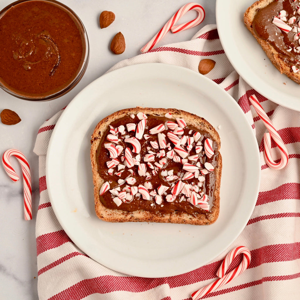 A slice of toast sits on a white kitchen plate the toast is covered in a layer of homemade chocolate almond peppermint butter and has crushed candy cane bits on top