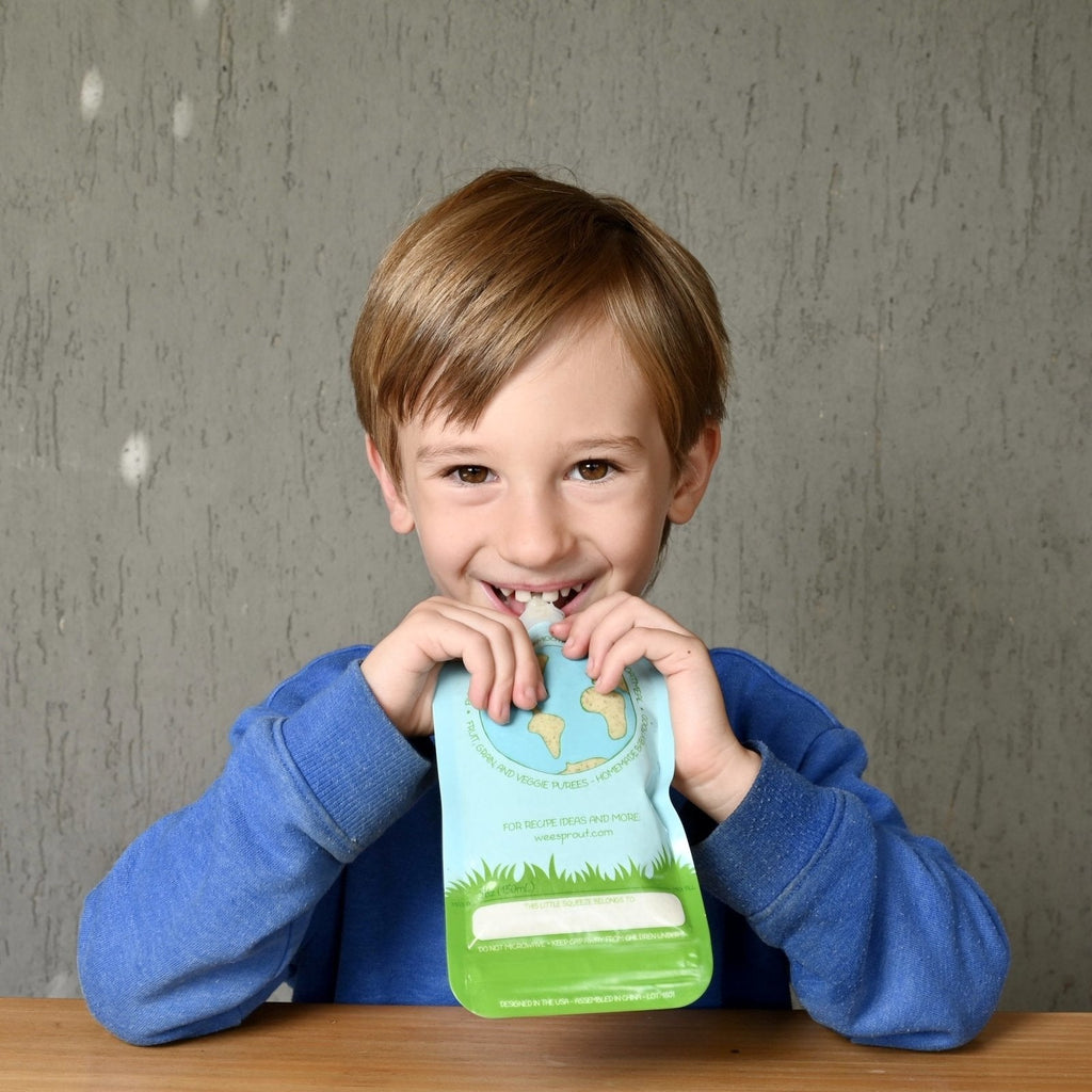 A little boy smiling at a kitchen table as he enjoys a fruit and vegetable pouch