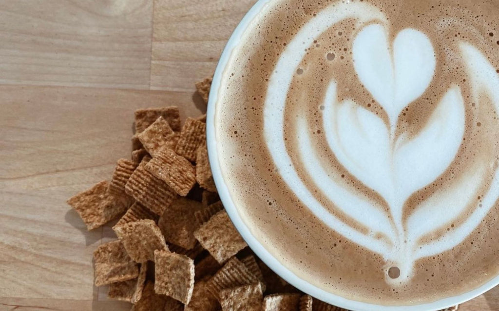 A coffee with latte art sits next to cinnamon toast crunch cereal on a wooden countertop.