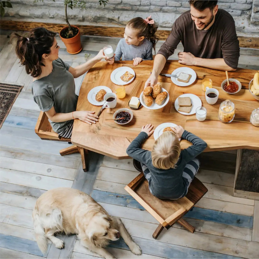 A family of four enjoying their breakfast. Their golden dog is sitting next to the table. The parents are drinking mugs of coffee.