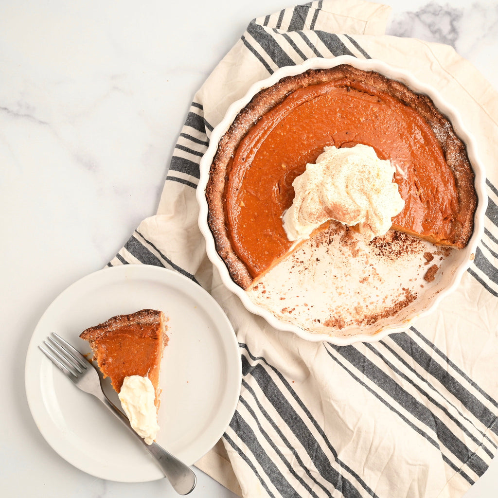 A white kitchen towel with blue stripes is set on a kitchen countertop with a pan of pumpkin pie resting on it with a small plate next to it with a piece of pumpkin pie and whipped cream on top