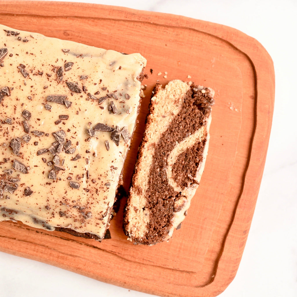 A wooden cutting board with a loaf of homemade marble cake resting on it with a sliced piece of the same marble cake cut off at the end of the loaf
