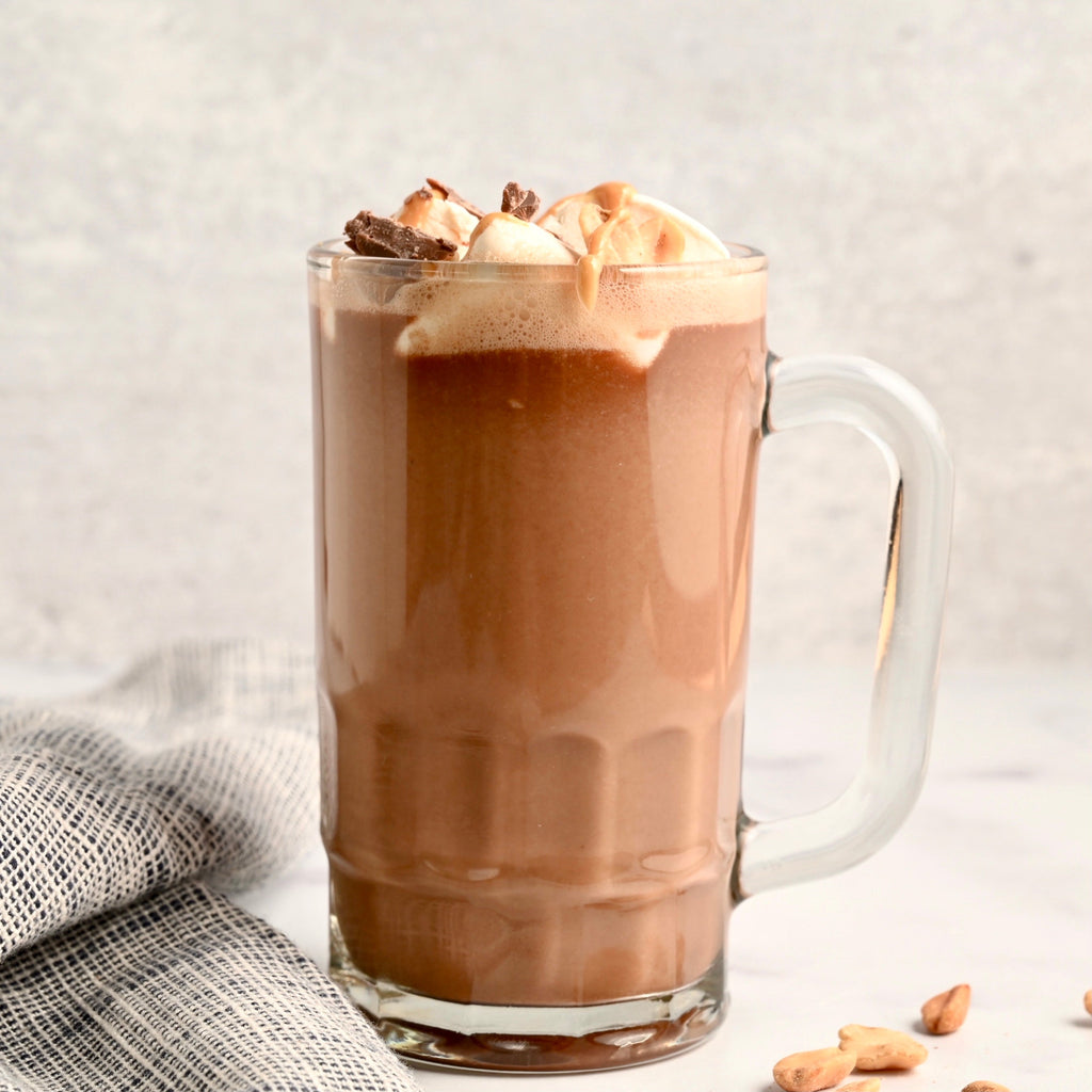 A large clear mug is full of hot chocolate and topped with roasted marshmallows and chocolate peanut butter candy pieces