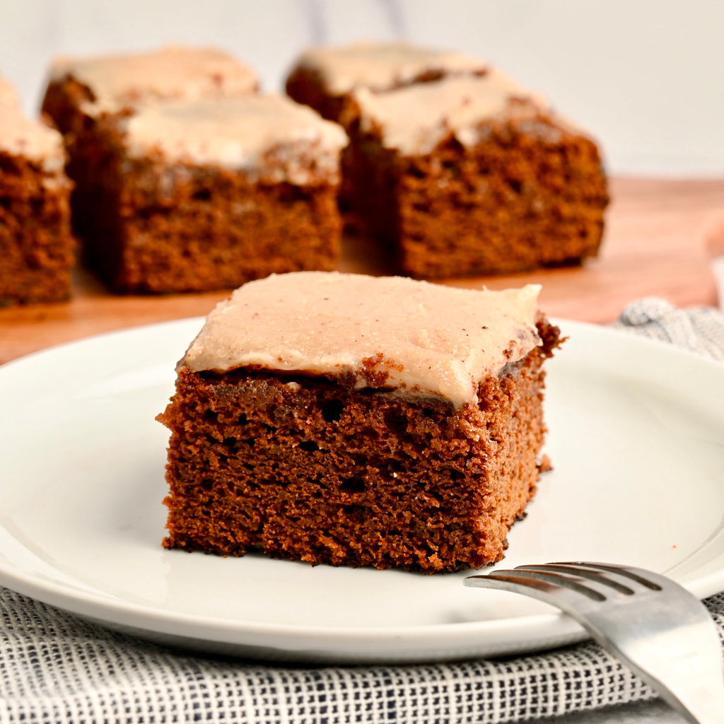A square slice of gingerbread cake is set upon a small white ceramic plate with a fork resting nearby