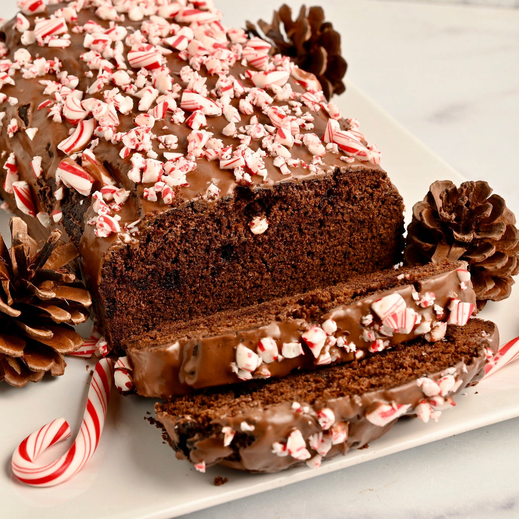 A chocolate loaf of cake is covered in crushed candy cane bits and is sliced on a white ceramic serving tray adorned with whole candy canes and pine cones