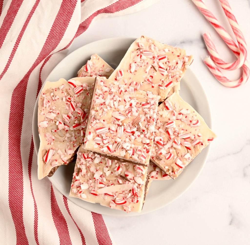 A white ceramic plate rests next to a white and red kitchen towel the plate has squares of homemade chocolate peanut butter peppermint bark stacked on it with whole candy canes resting near the plate