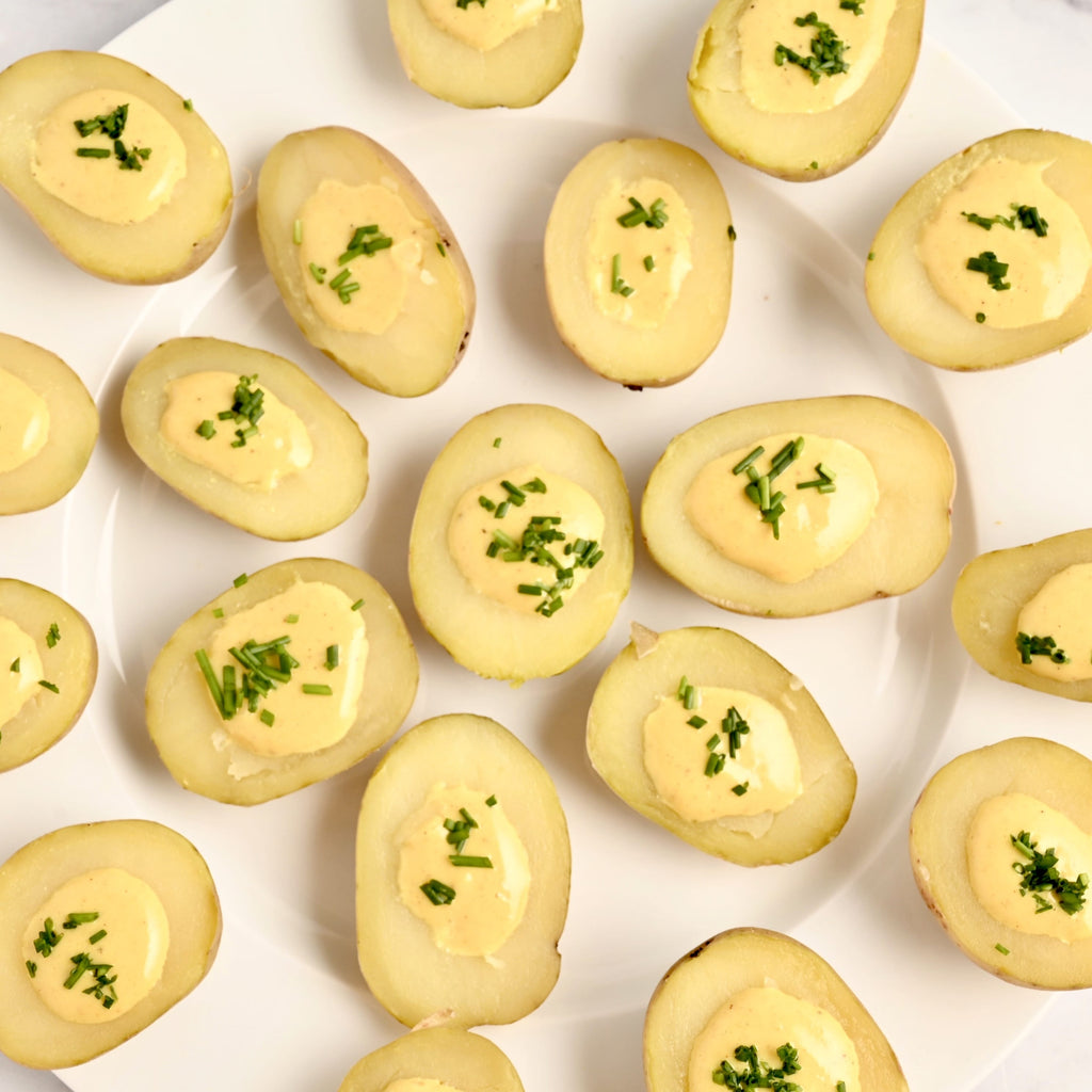 A white plate covered in bite sized potatoes each filled with a savory cashew sauce and topped with fresh green herbs for garnish