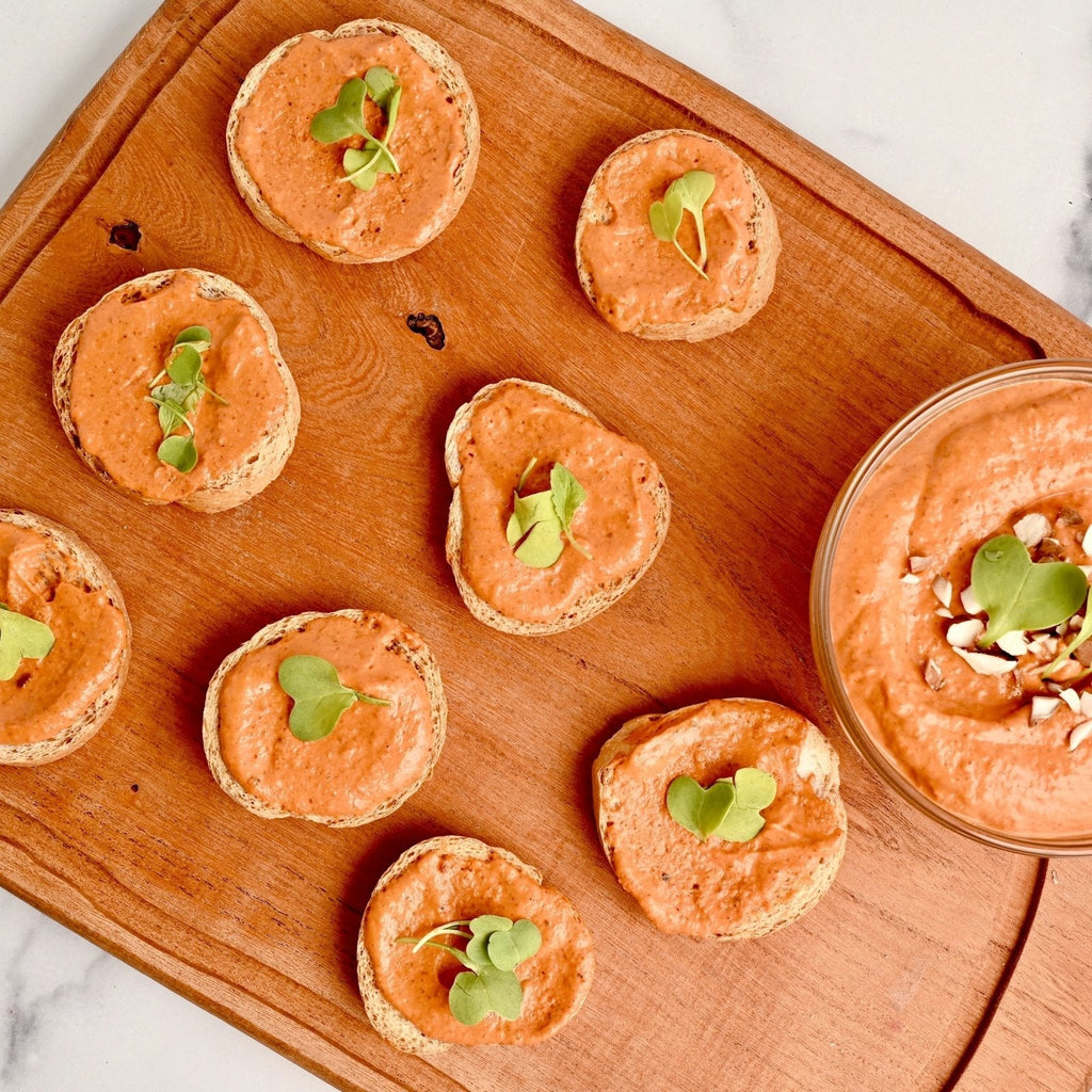 A wooden cutting board covered in small circles of bread covered in almond butter romesco spread and a few sprigs of micro greens