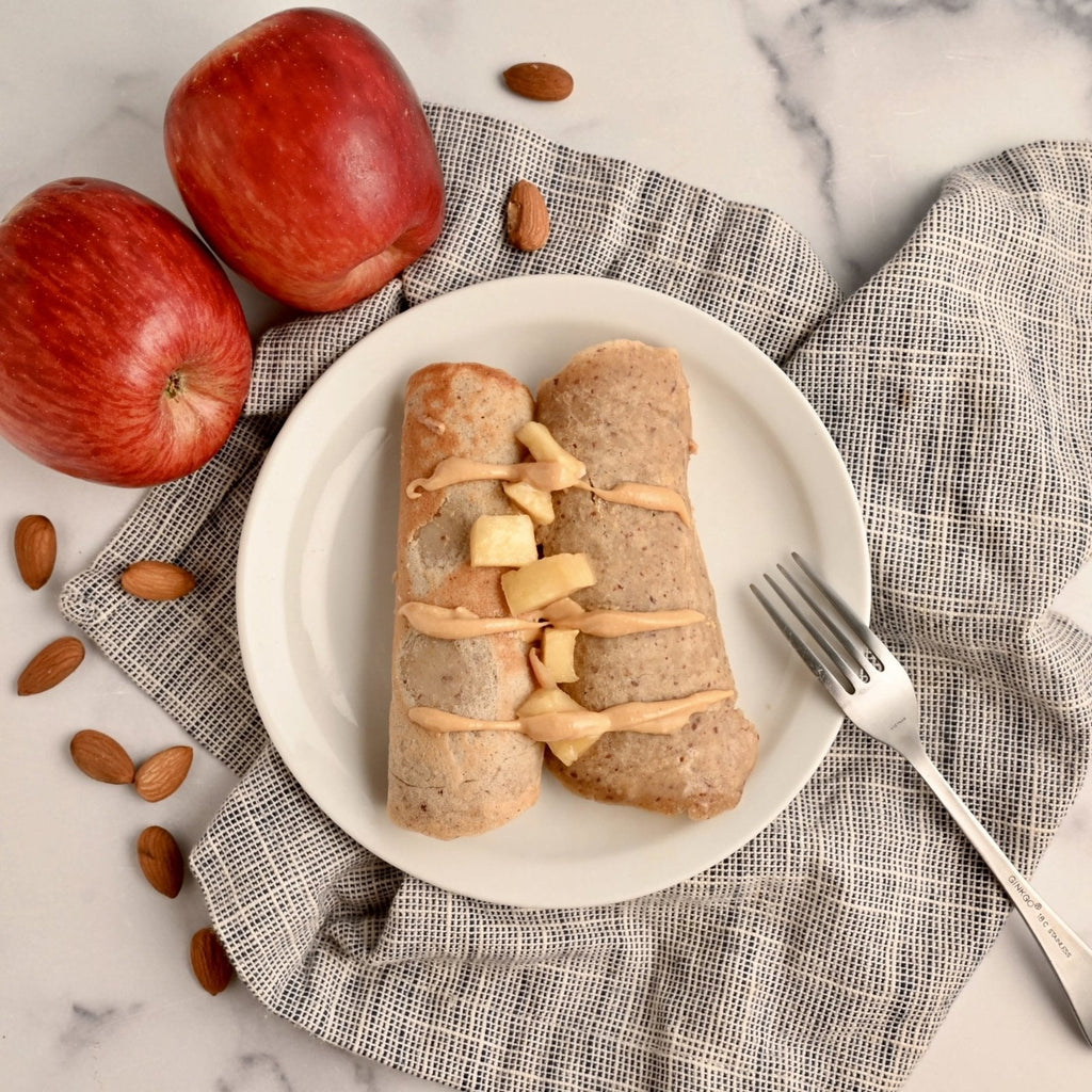 White plate with two apple cinnamon crepes topped with homemade nut butter with some whole red apples off to the side with a few almonds