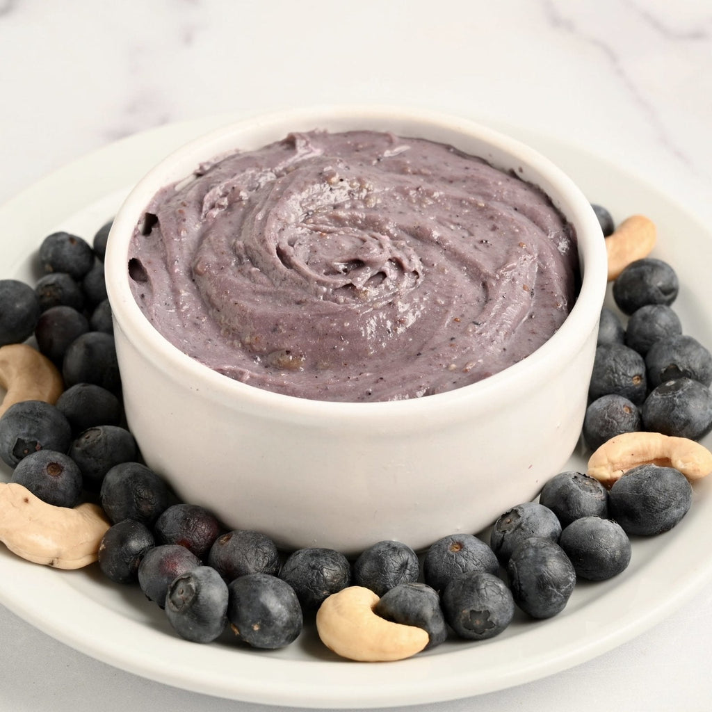 White plate with a white bowl in the center filled with homemade blueberry cream cheese and the plate is covered in fresh blueberries and cashews