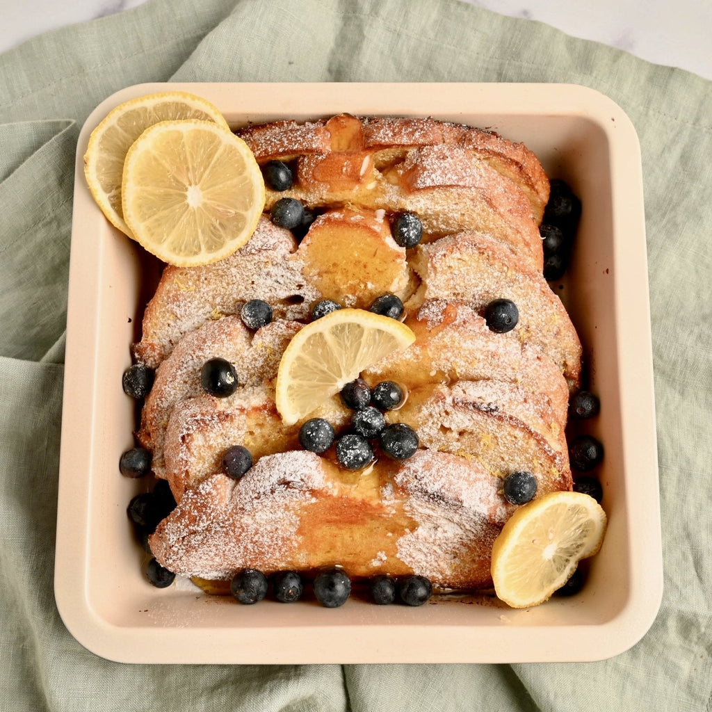 Tan casserole dish filled with slices of homemade French toast and topped with blueberries and slices of fresh lemon
