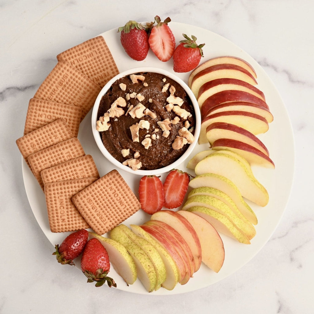 A white plate covered in crackers and fresh fruit slices with a small white bowl in the center that is filled with brownie batter dip and topped with crushed walnut pieces