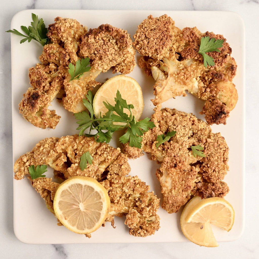 A white square plate covered in crispy crunchy cauliflower steaks along with fresh lemon slices and fresh parsley sprigs