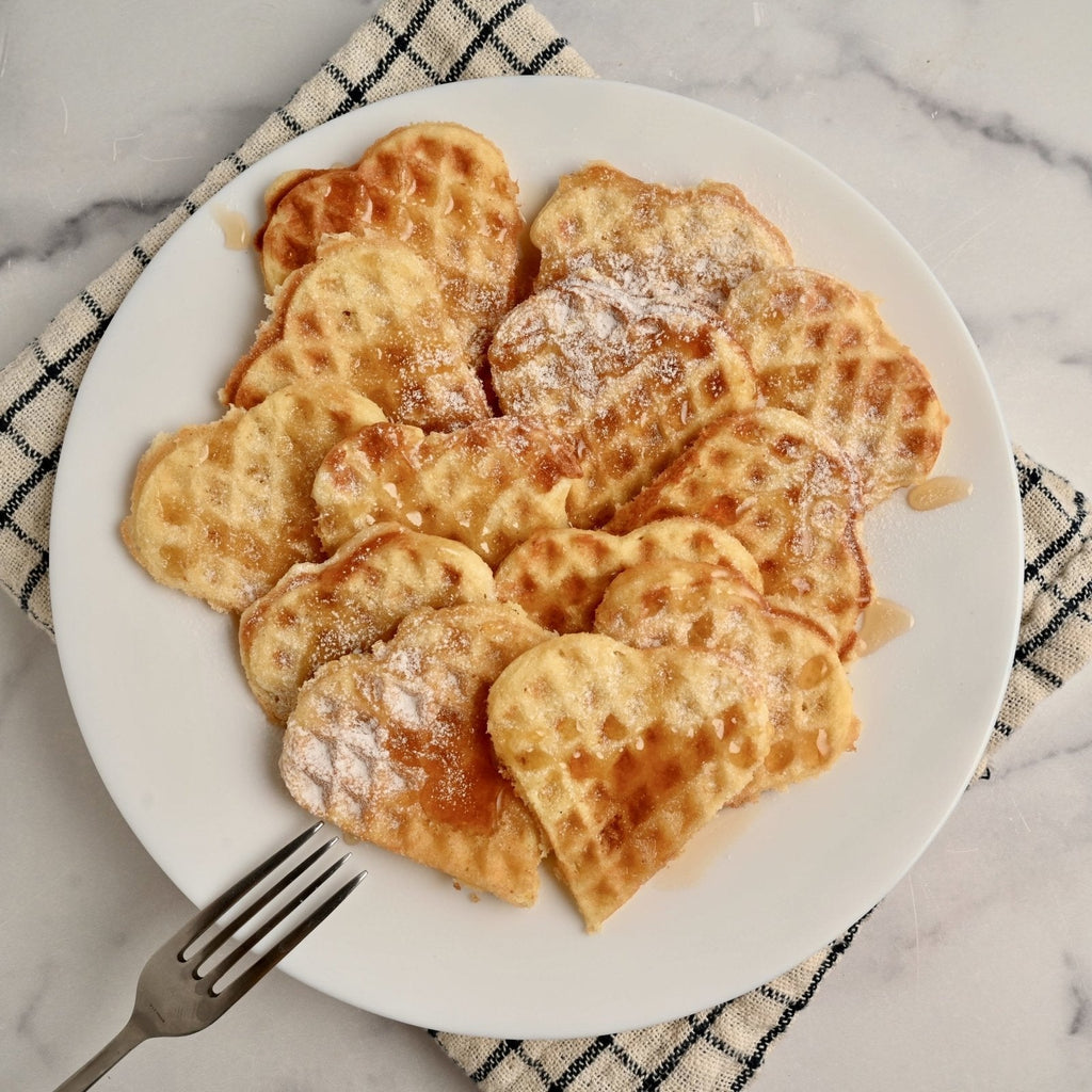 A white ceramic plate with mini heart-shaped waffles covered in syrup and powdered sugar