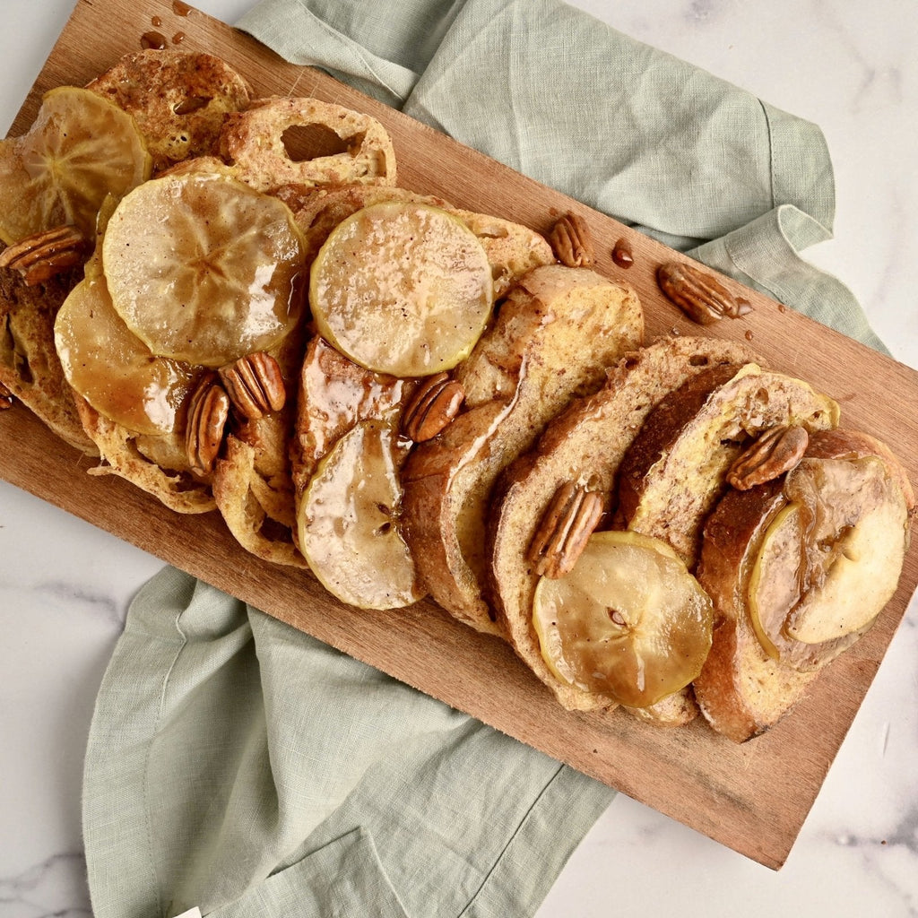 A wood cutting board covered in French toast slices that have been topped with apple slices