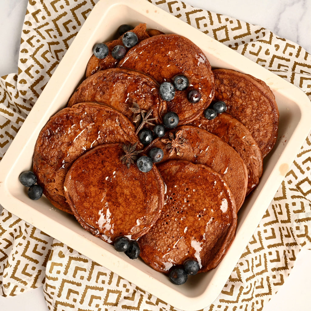 A large white serving dish is stacked layers deep with homemade gingerbread pancakes topped with a drizzle of maple syrup and a handful of fresh blueberries