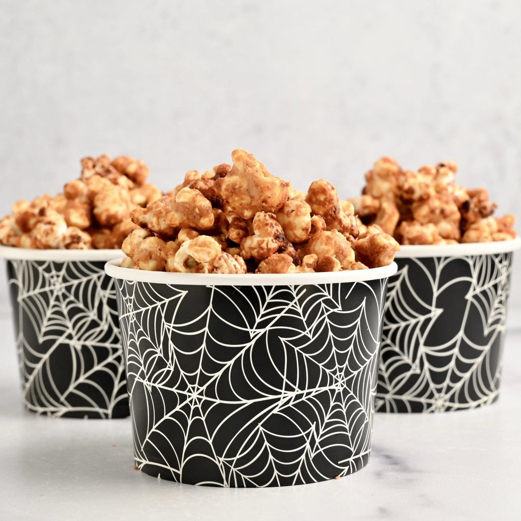 Three black paper cups with white spider webs on them with each cup filled with nut butter popcorn