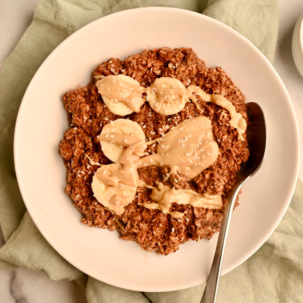 A white bowl of chocolate overnight oats with banana slices on top and nut butter