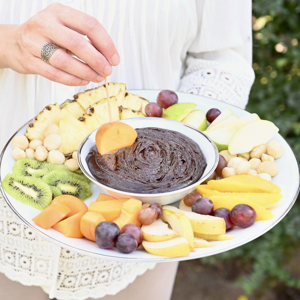 A white ceramic platter covered in fresh fruit with a bowl of chocolate and macadamia nut fondue in the center for dipping