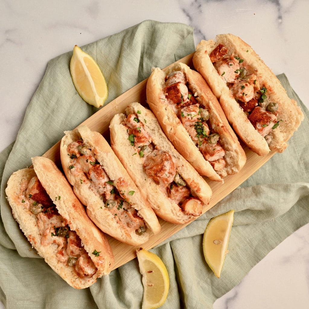 A long wooden cutting board with five homemade lobster rolls laid across it with lemon wedges of to the sides