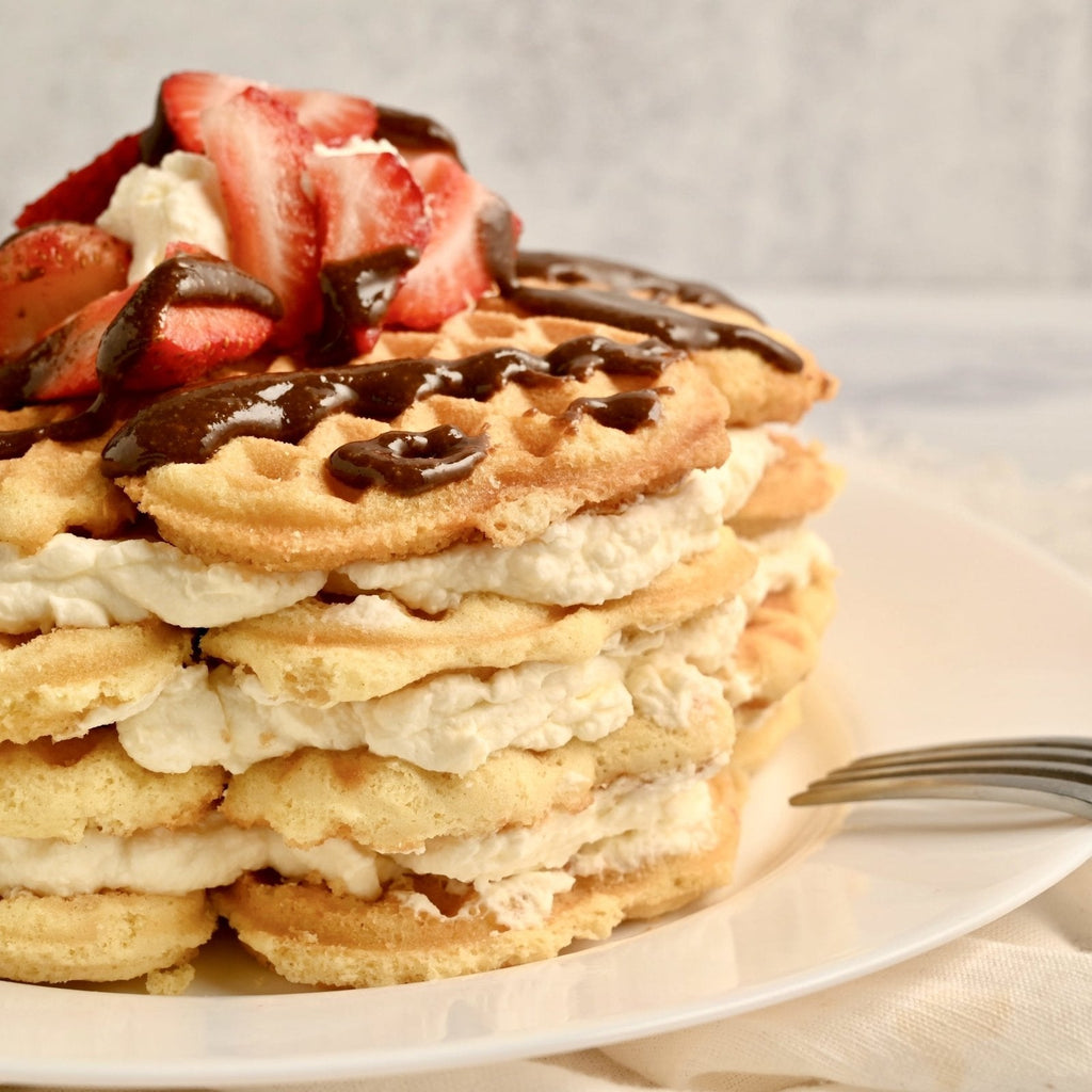 Waffles stacked to make a cake with coconut whipped cream in between each waffles and chocolate nut butter  drizzled on top with slices of fresh strawberries