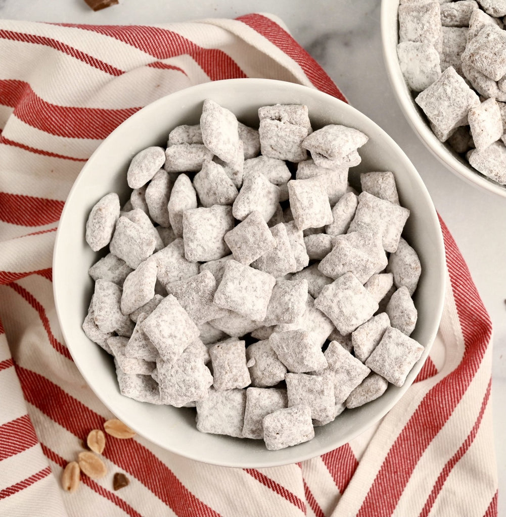 A red and white striped kitchen towel lays across a kitchen counter and on top of the towel is a white ceramic bowl filled with homemade puppy chow dusted in powdered sugar