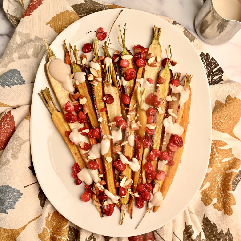 A white platter of roasted carrots with bright red cranberries on top with a drizzle of sauce