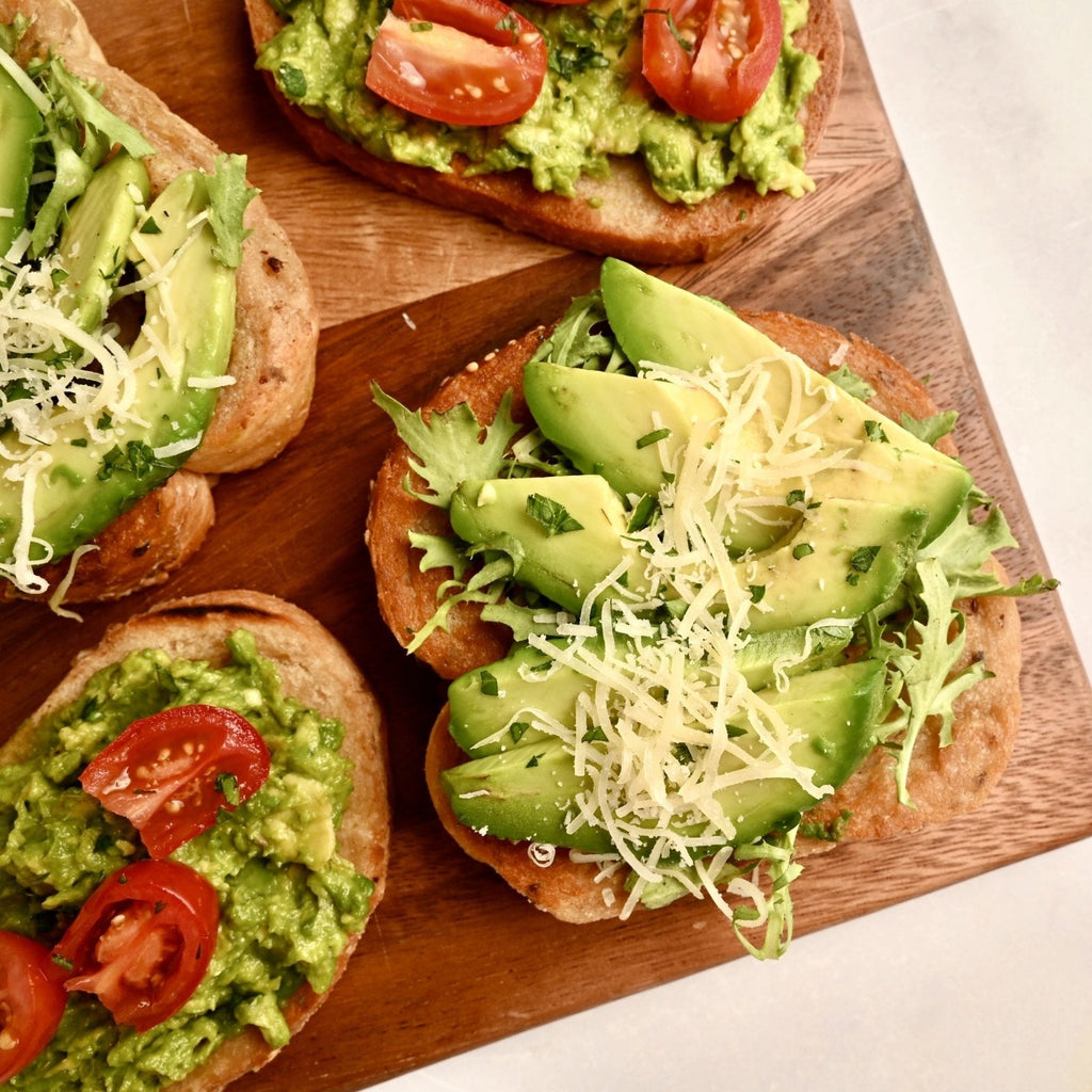 A wooden cutting board with thick slices of bread topped with savory ingredients like avocado slices, sprouts, and tomatoes