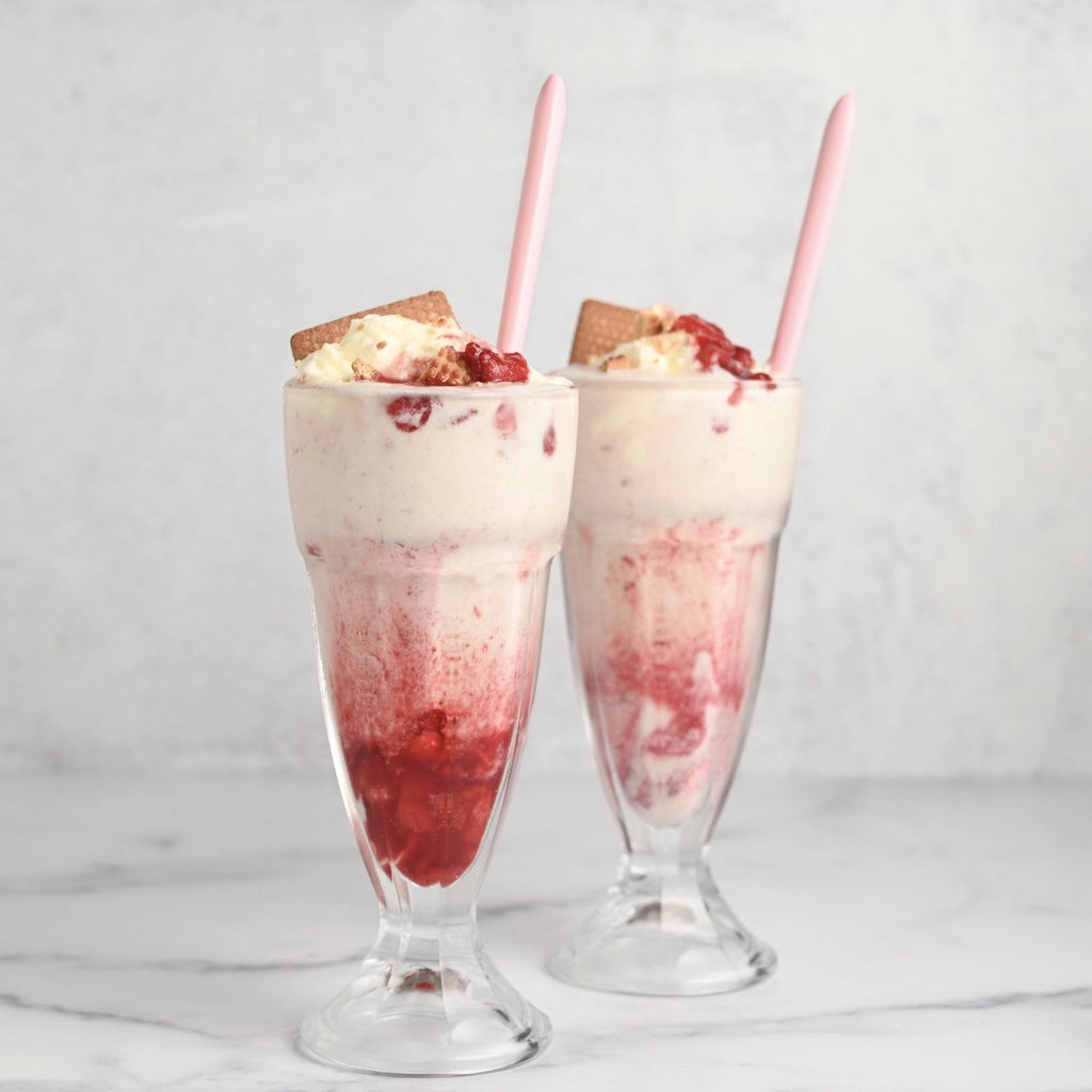 Two clear milkshake glasses each filled with a bright pink strawberry milkshake and topped with crumbled graham crackers and a straw