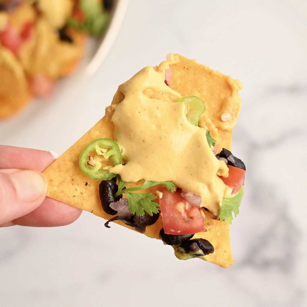 A close up image of one tortilla chip covered in creamy cheese sauce, black olives, jalapeño pepper slices and cubed tomatoes