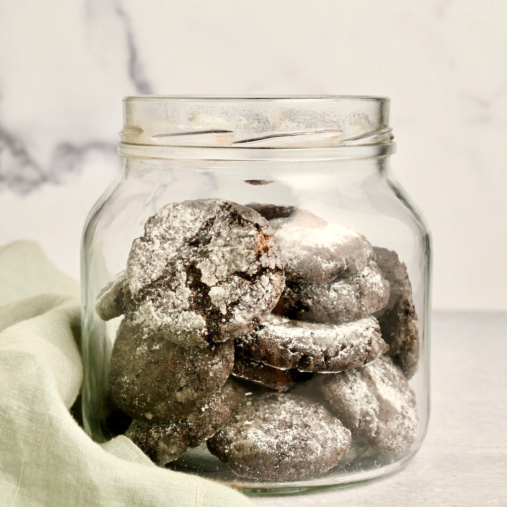 A clear glass jar sits next to a light green kitchen towel and the jar is filled with dark chocolate cookies that have been dusted with white powdered sugar