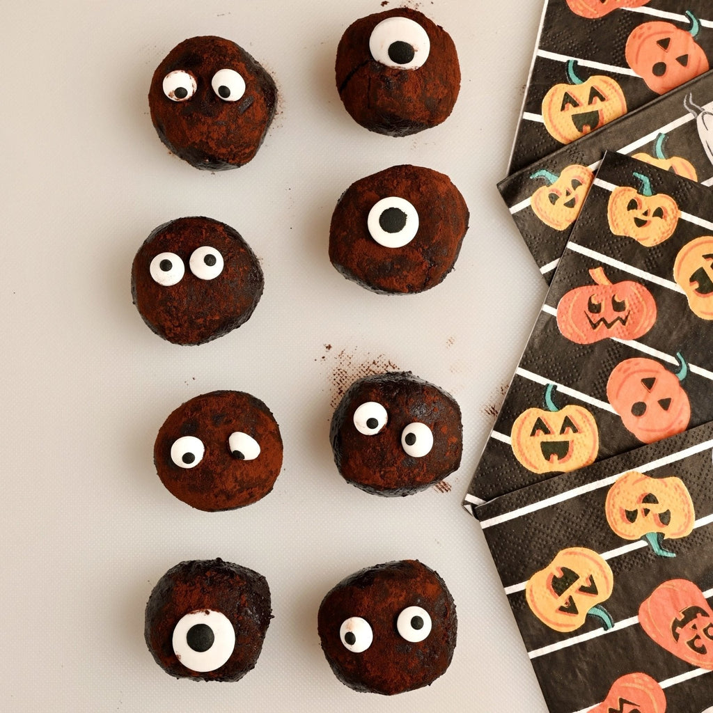 White background with chocolate truffle balls with eyes on each black napkins covered in orange pumpkins are off to the side