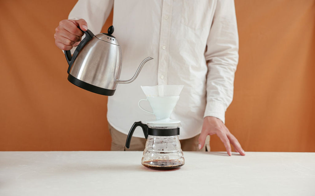 A man is pouring water from a Bonavita kettle for pour over coffee in front of an orange background.