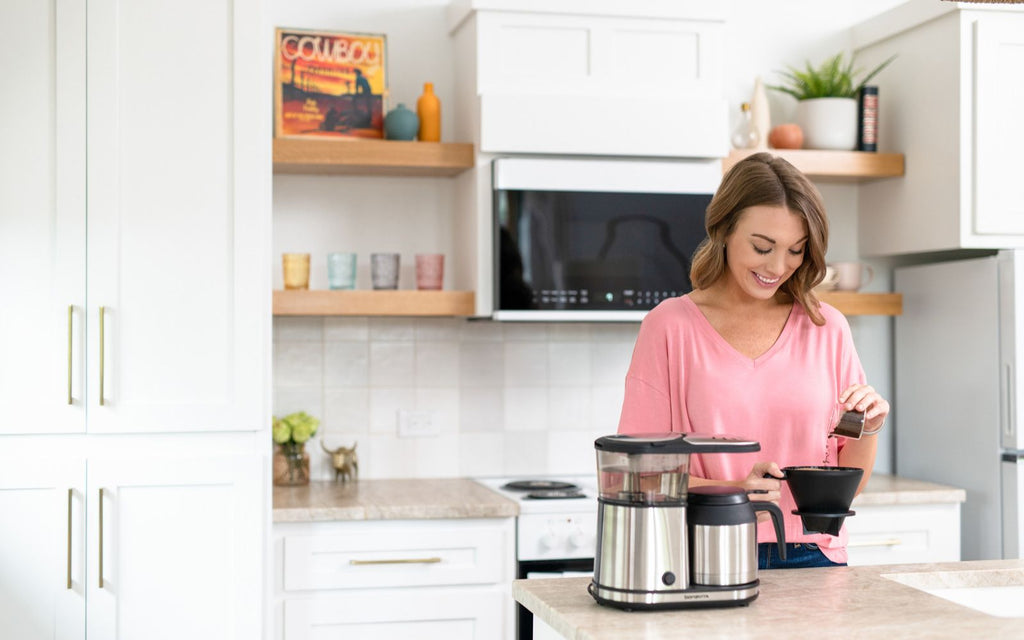 A woman is pouring coffee grinds into the Connoisseur Coffee Brewer in her minimalist kitchen.