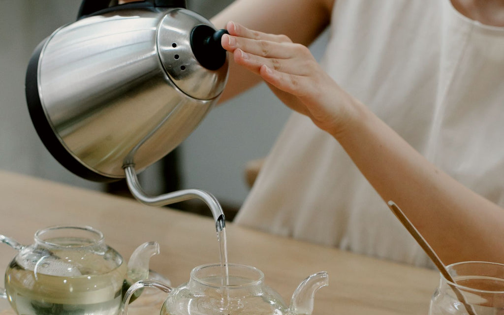 A woman is pouring water from a Bonavita kettle into a glass teapot.