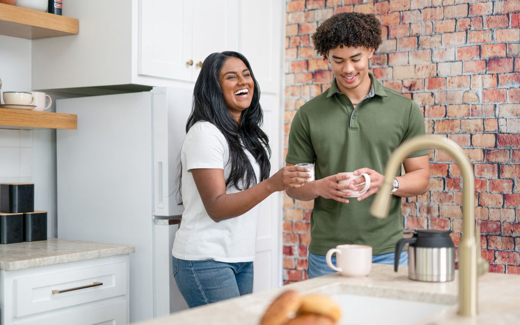 A woman and man are in a kitchen with mugs in their hands, laughing over drinking coffee.