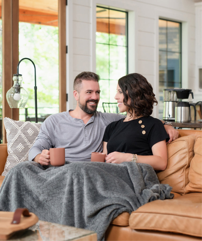 A young couple sitting on a sofa enjoying a cup of coffee. They have a blanket over their knees.