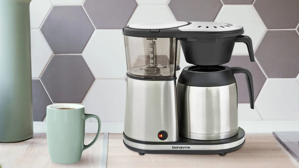 A Bonavita coffee brewer is placed on a kitchen counter. Grey and white hexagonal tiles are featured in the background with a pale green coffee mug is to the left of the coffee brewer.  